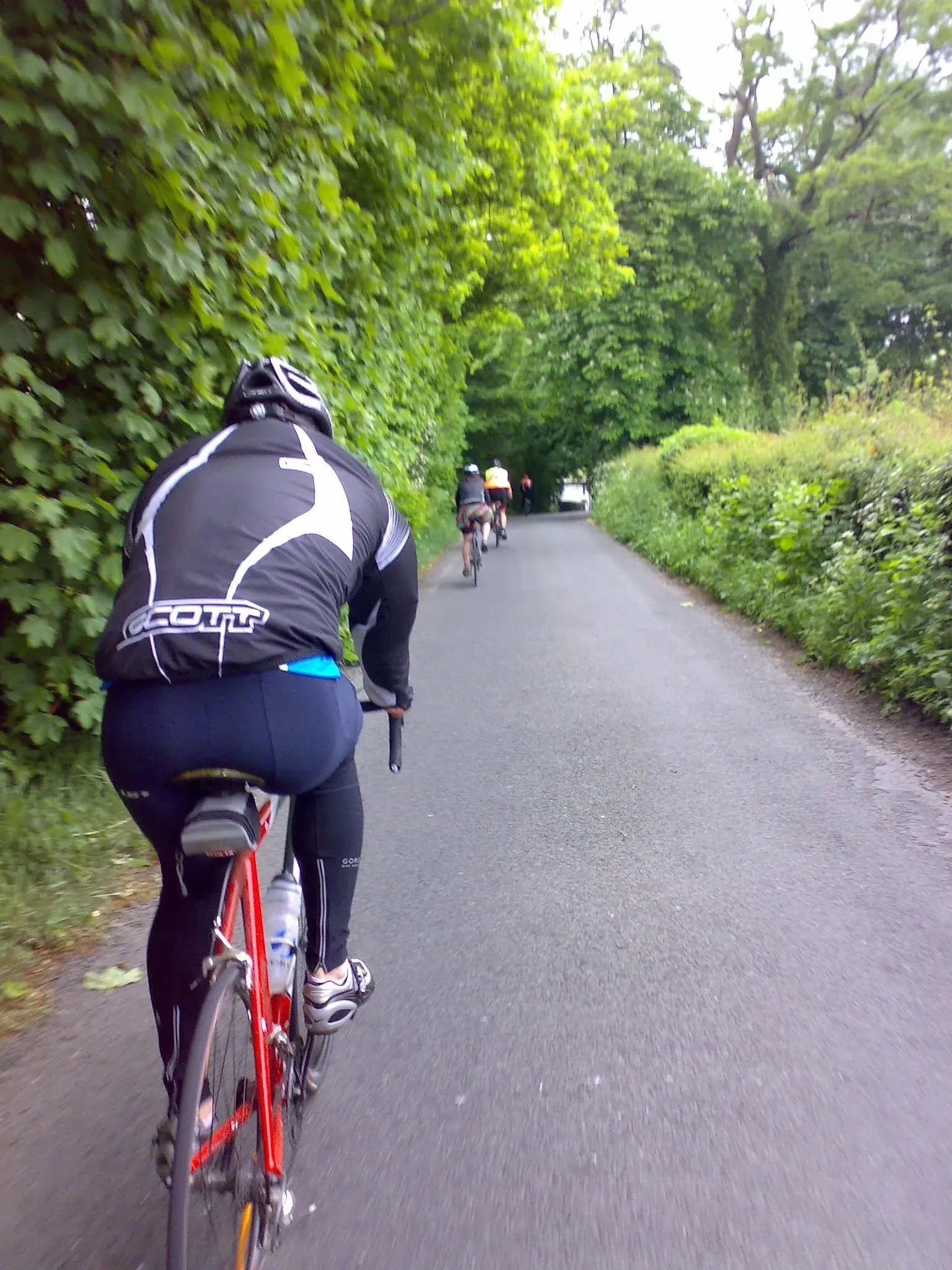 Cyclist in black gear riding red bike on tree-lined country lane