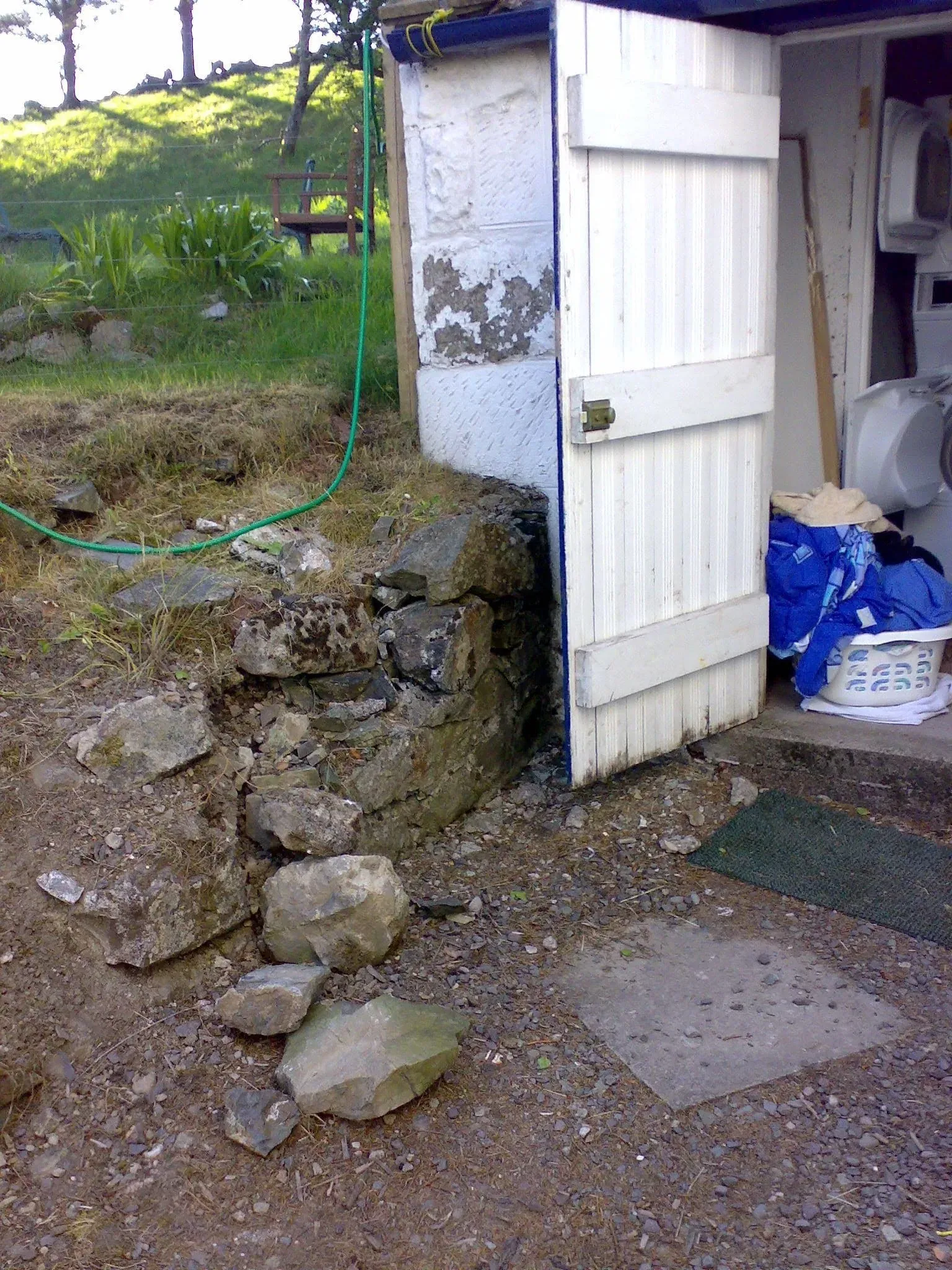 Simple white shed structure with green hose and stone foundation wall