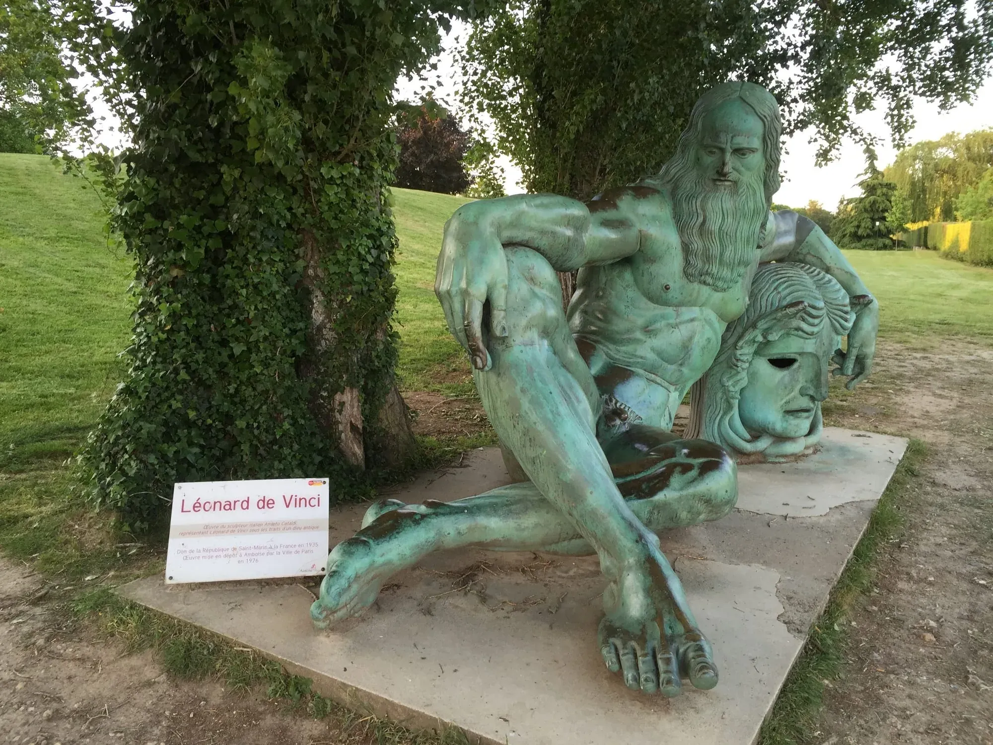 Green copper sculpture of reclining bearded man labeled Léonard de Vinci in park setting