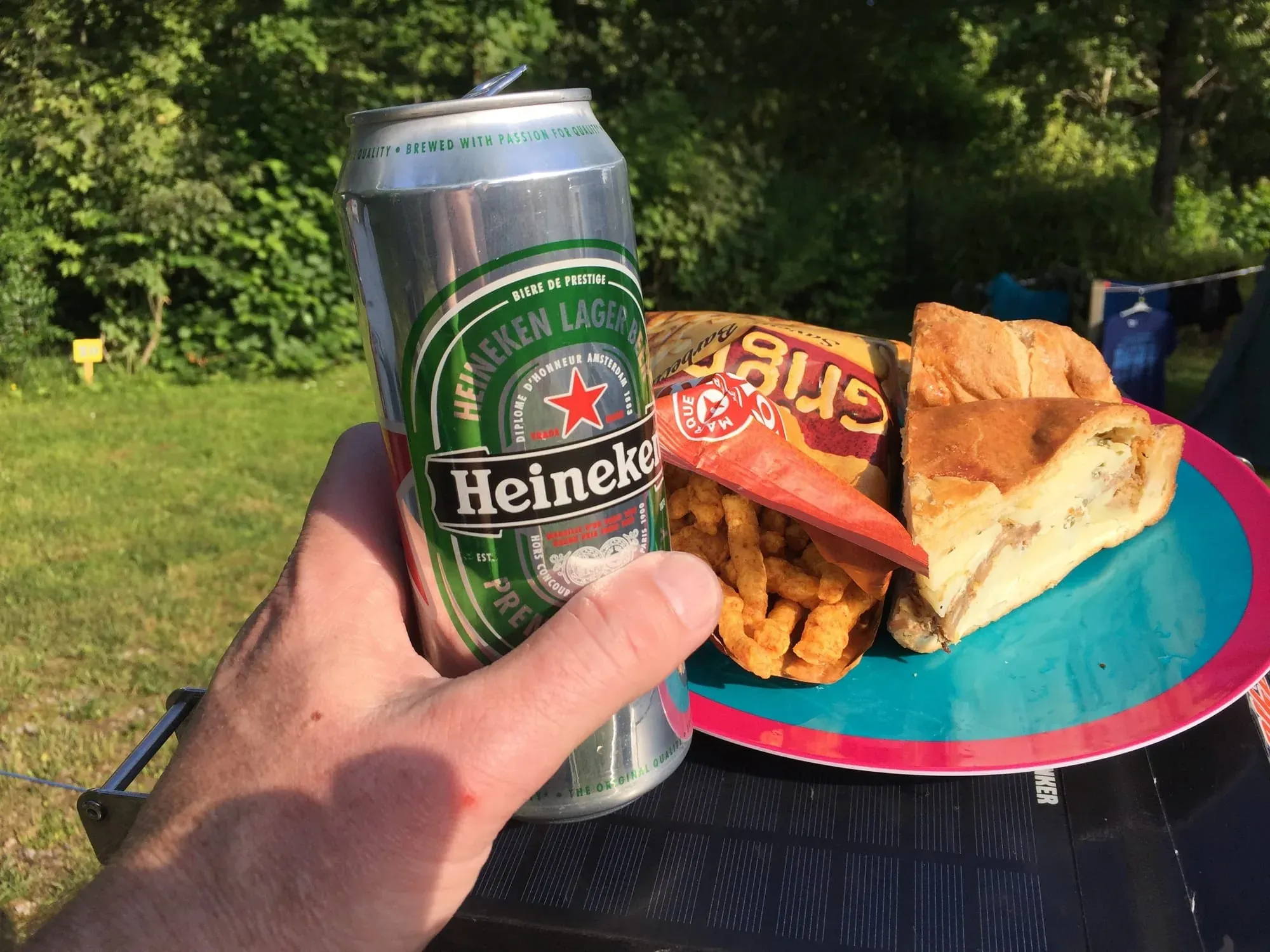 Hand holding Heineken beer can with sandwich and chips outdoors in grass setting