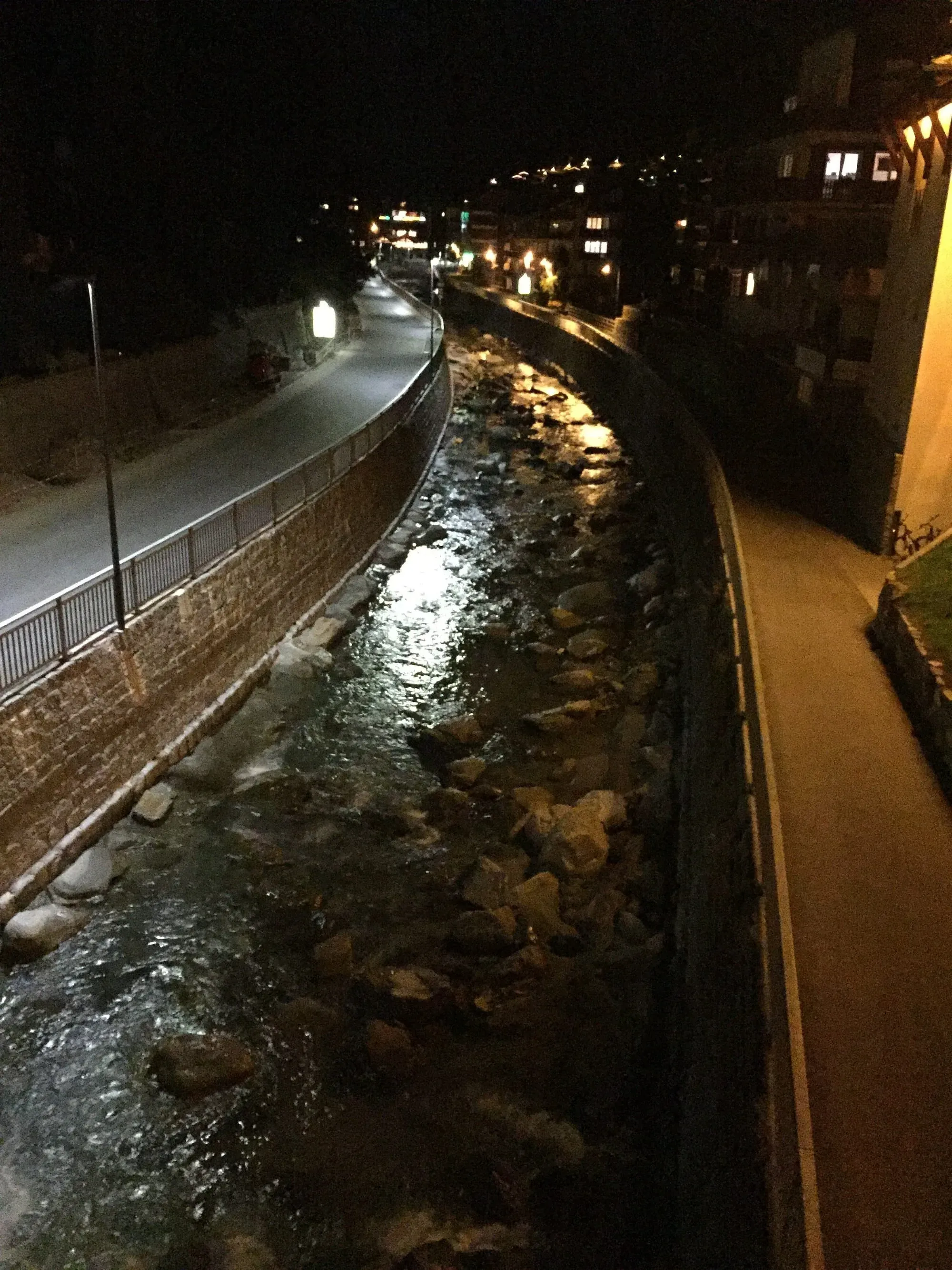 Night view of illuminated pathway alongside stream with buildings and mountains