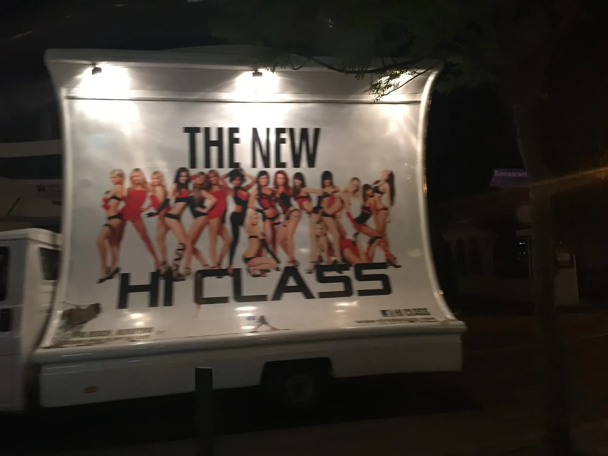 Illuminated billboard advertising "The New Hi Class" with group of women in red and black outfits