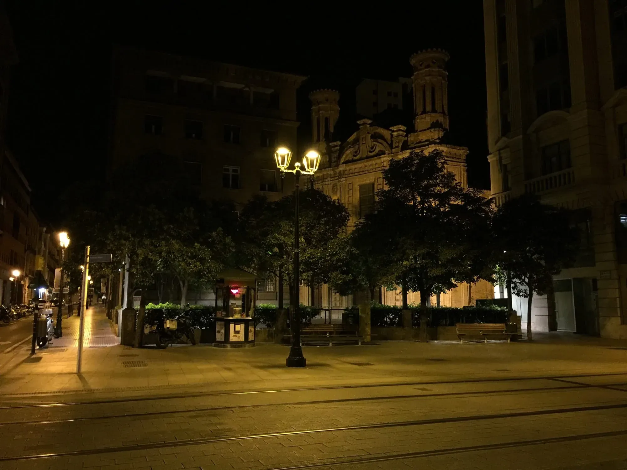 Nighttime plaza with illuminated church building and street lamps, tram tracks visible