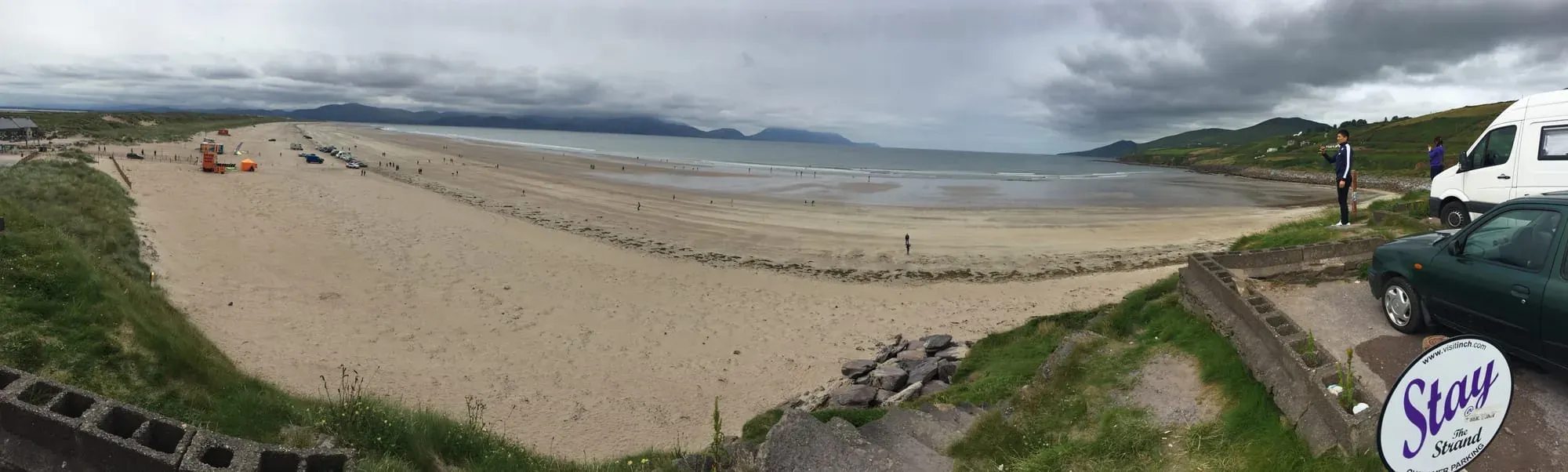 Sandy beach with mountains across bay, parked vehicles and Stay Strand sign visible