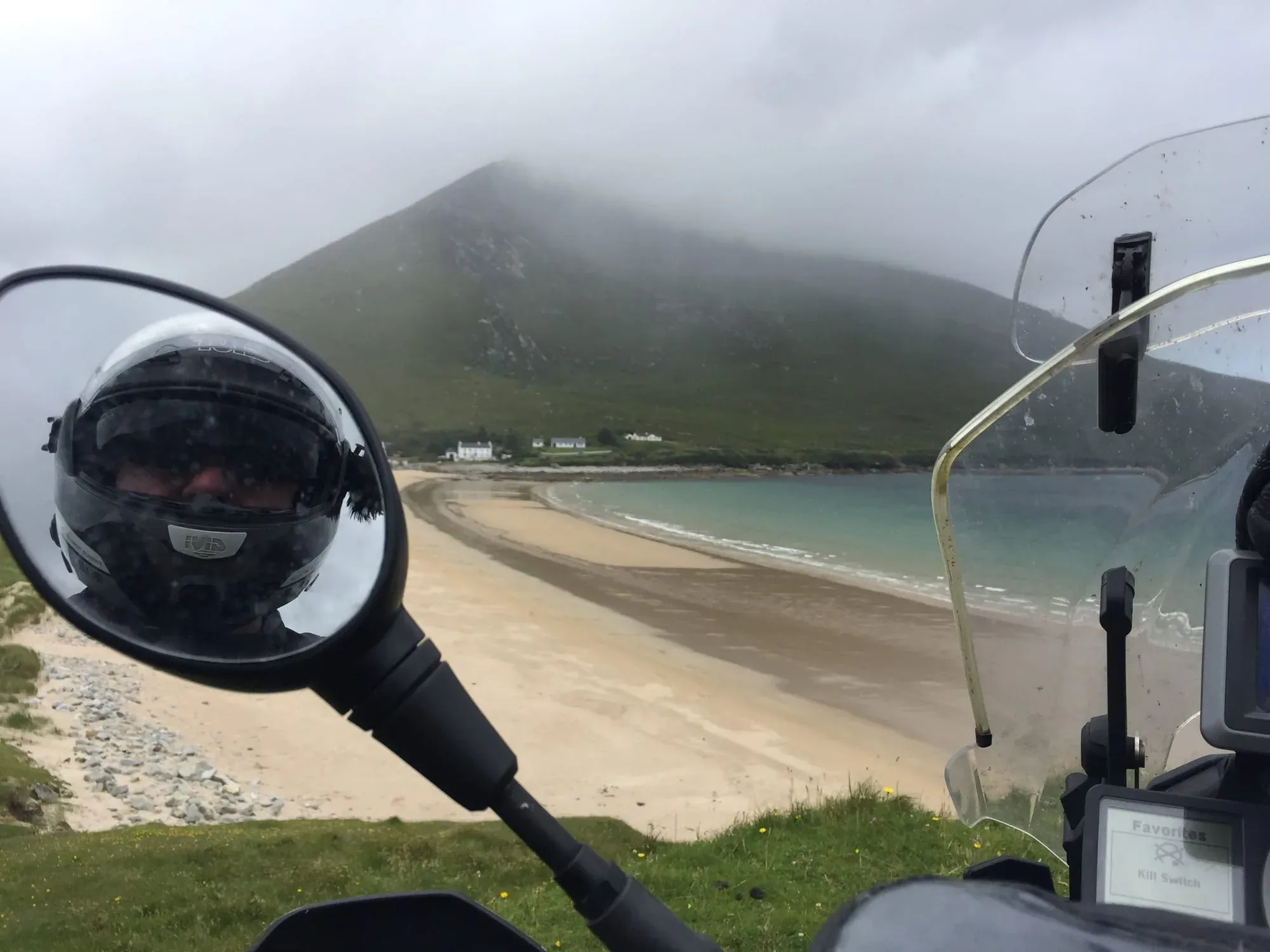 Motorcycle mirror reflection of helmet and beach cove with misty mountain backdrop