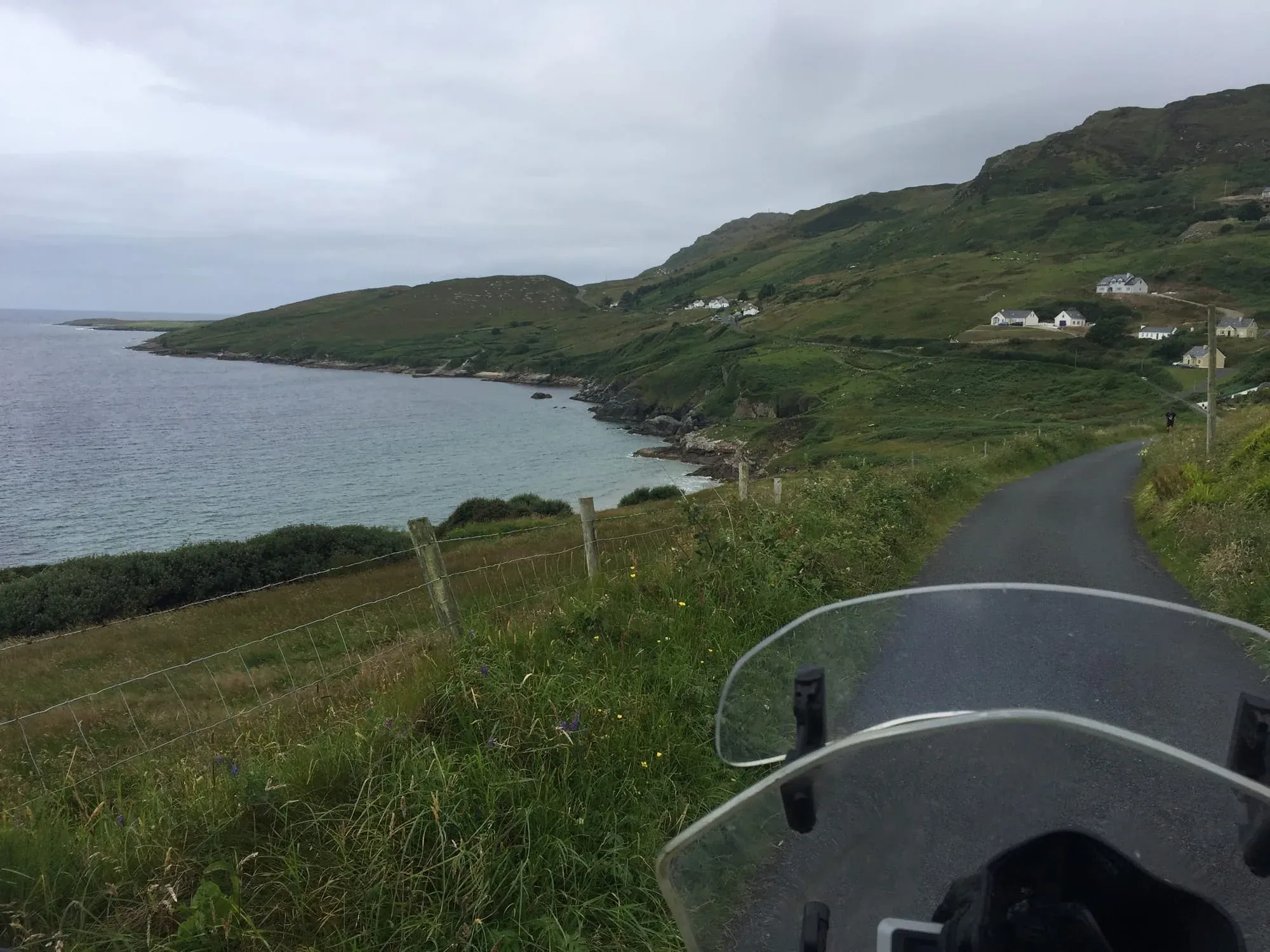 Motorcycle handlebar view of coastal road, green cliffs, and small white houses