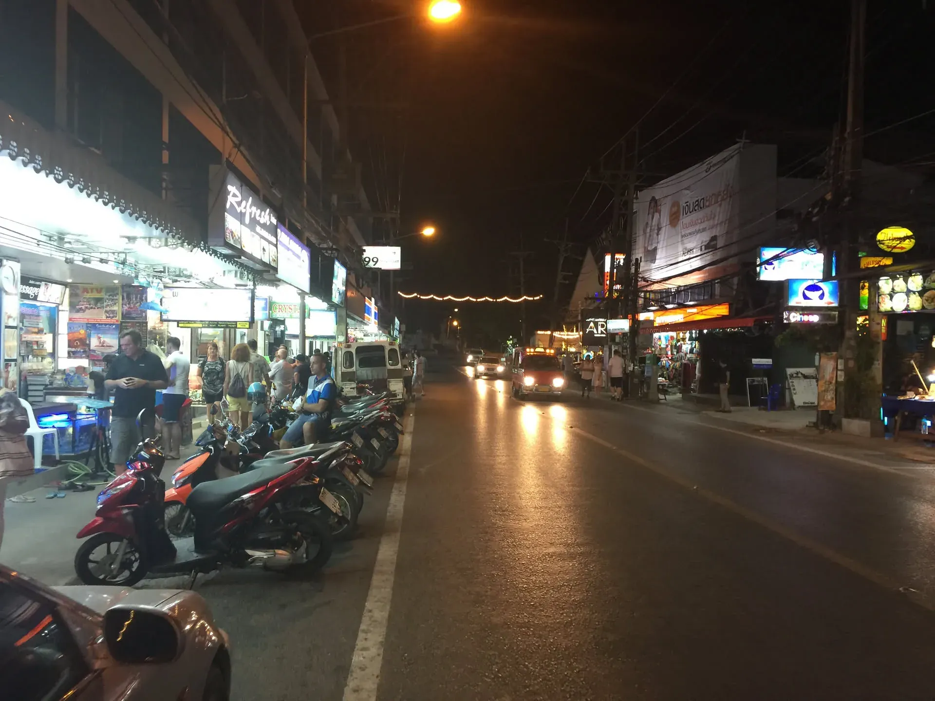 Wet night street in Karon with scooters and shops