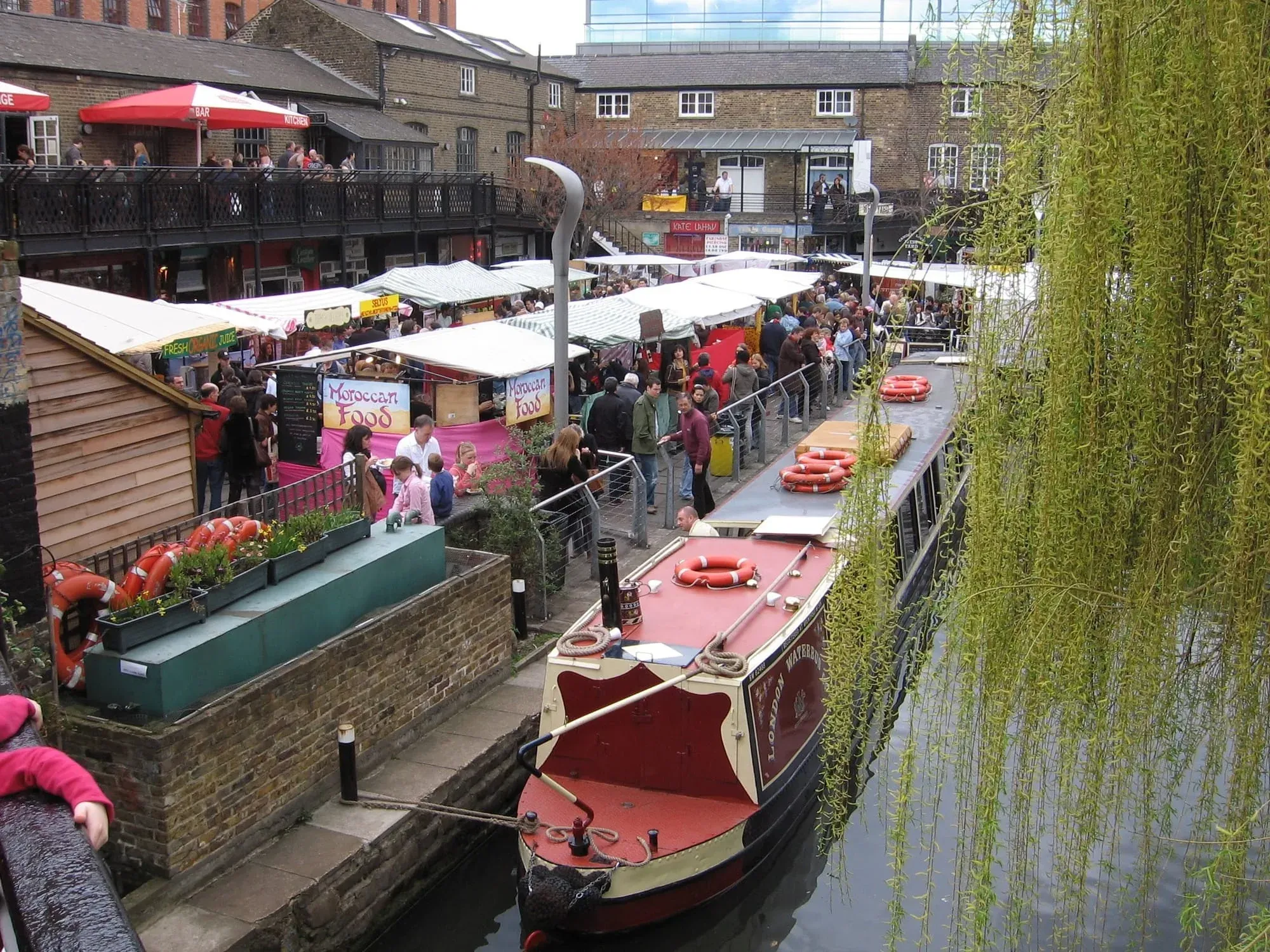 Aerial view of busy canal-side market with food stalls, crowds, and moored boats