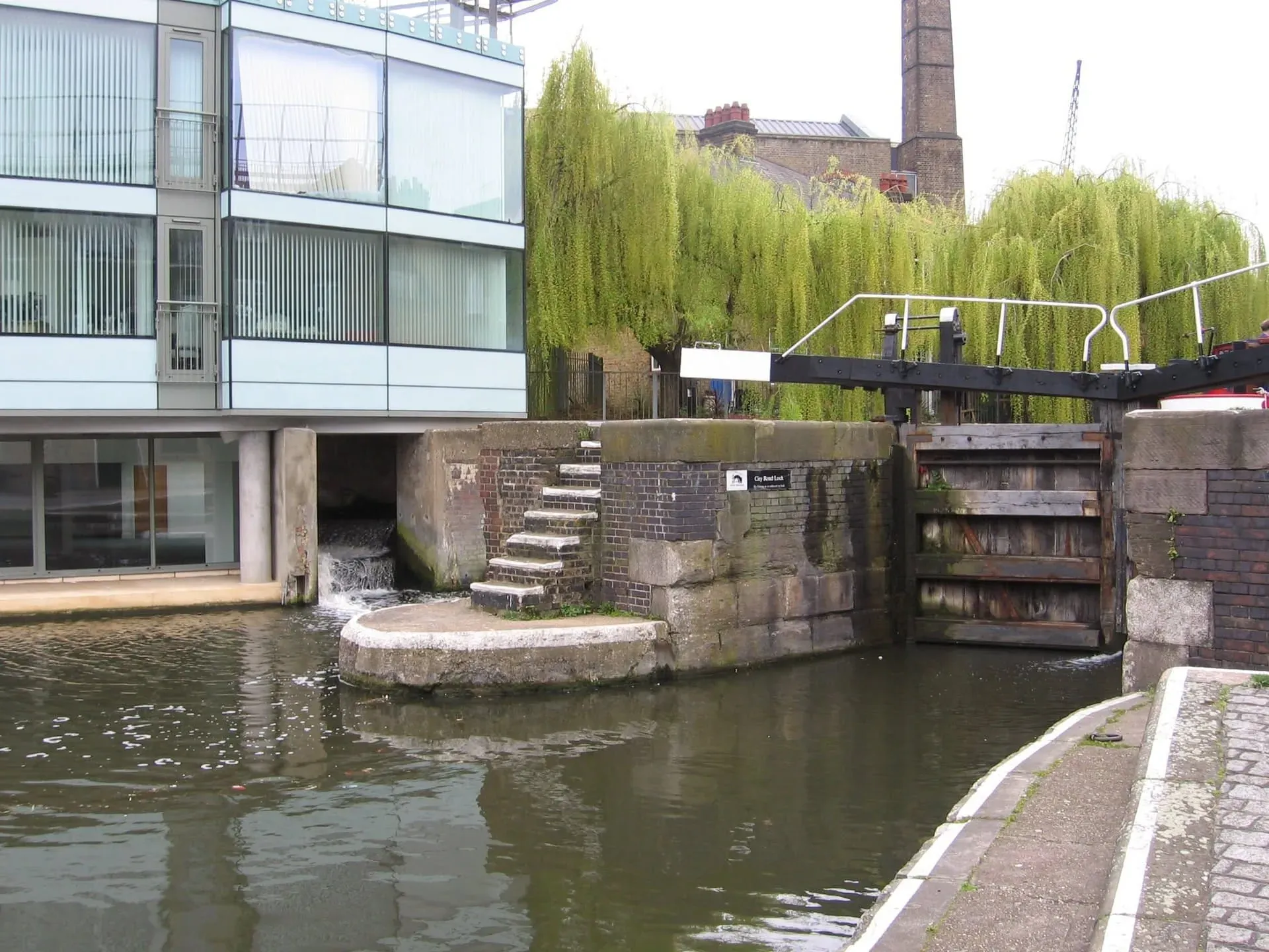 Canal lock mechanism with modern building and willow trees in background