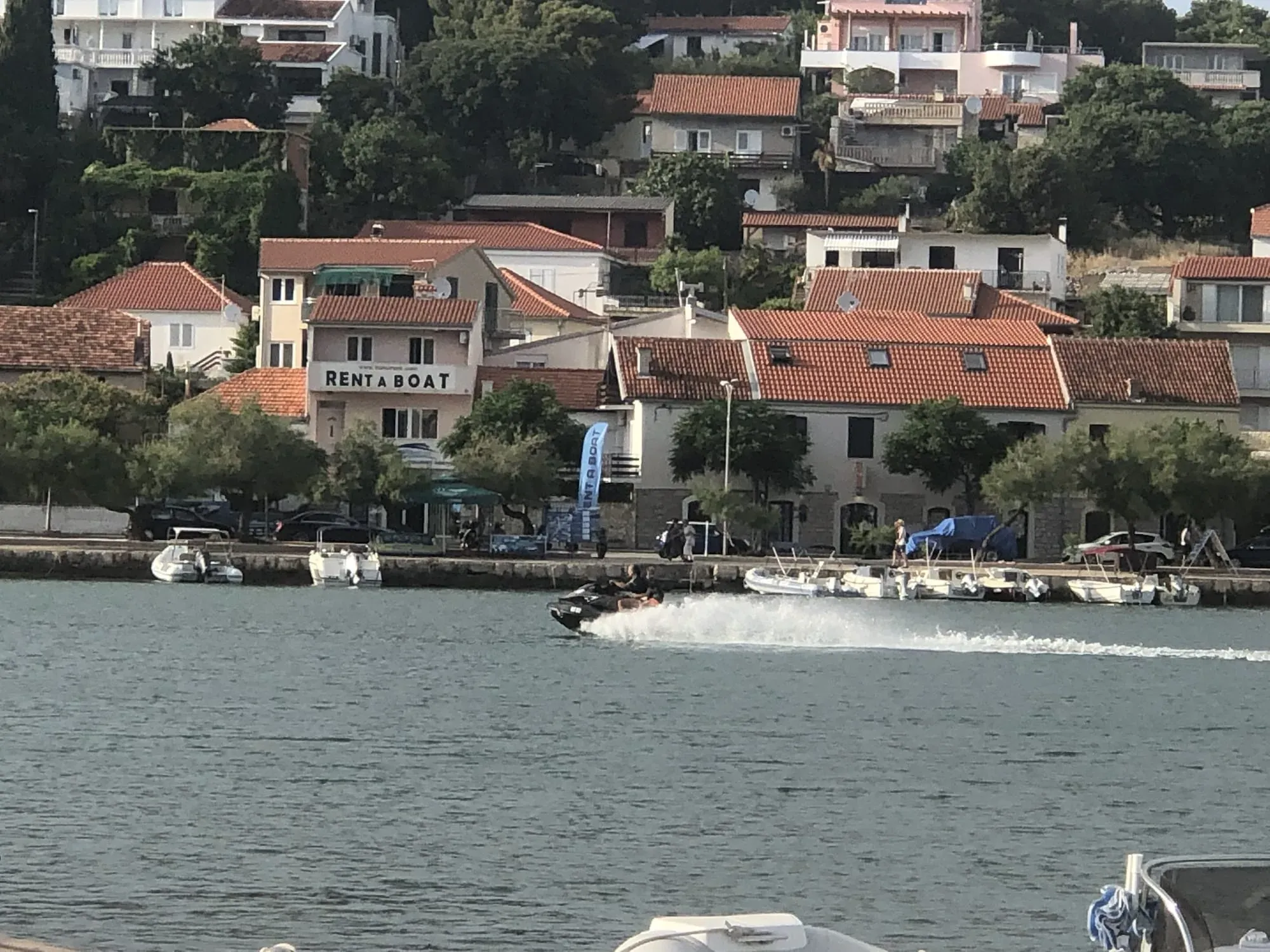 Coastal town with red-roofed buildings, boat rental shop, and moored boats in harbor