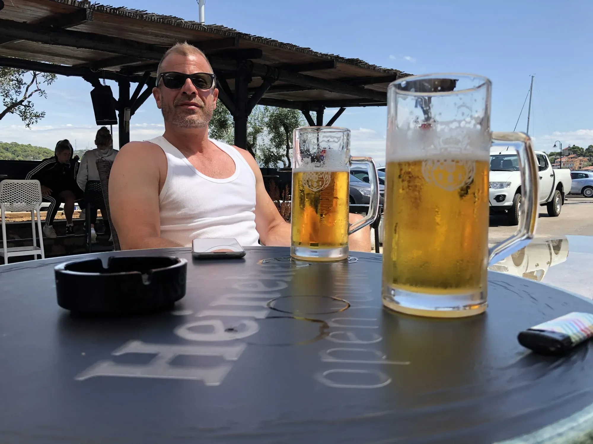 Man in white tank top sitting at outdoor table with beer glasses in daylight