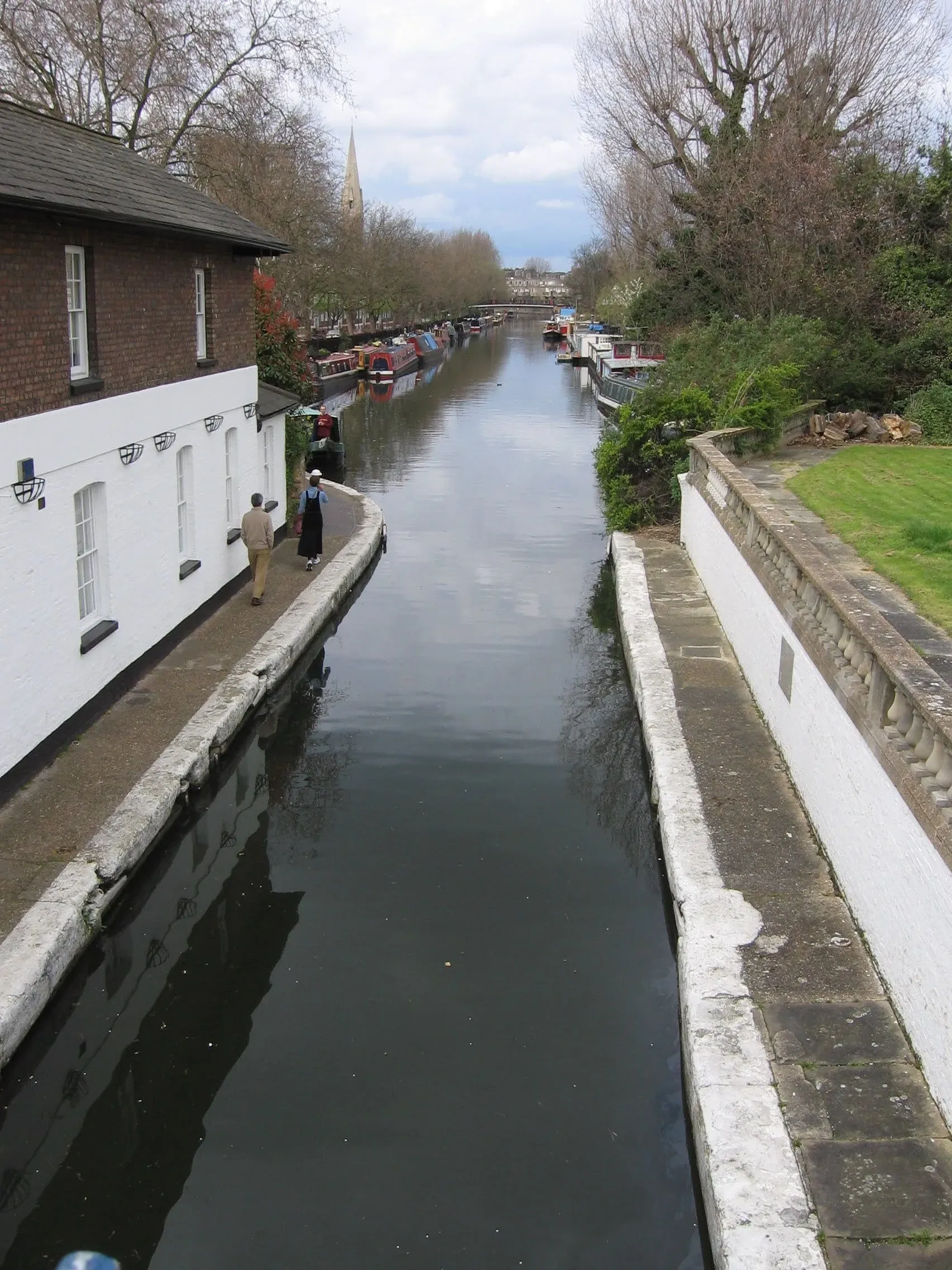 Narrow canal with moored boats between white buildings and trees
