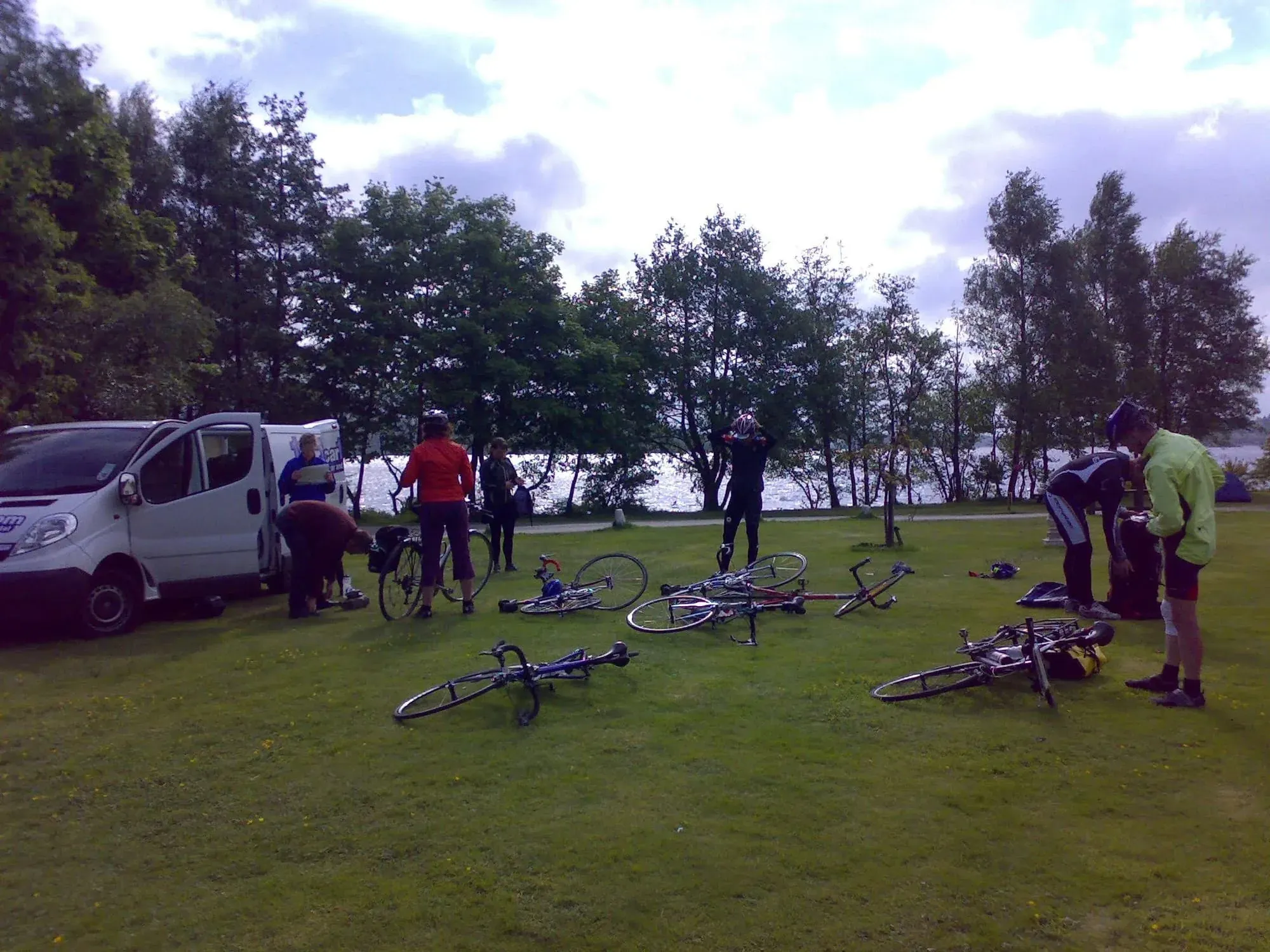 Multiple bicycles on grass by lakeshore with van and cyclists preparing
