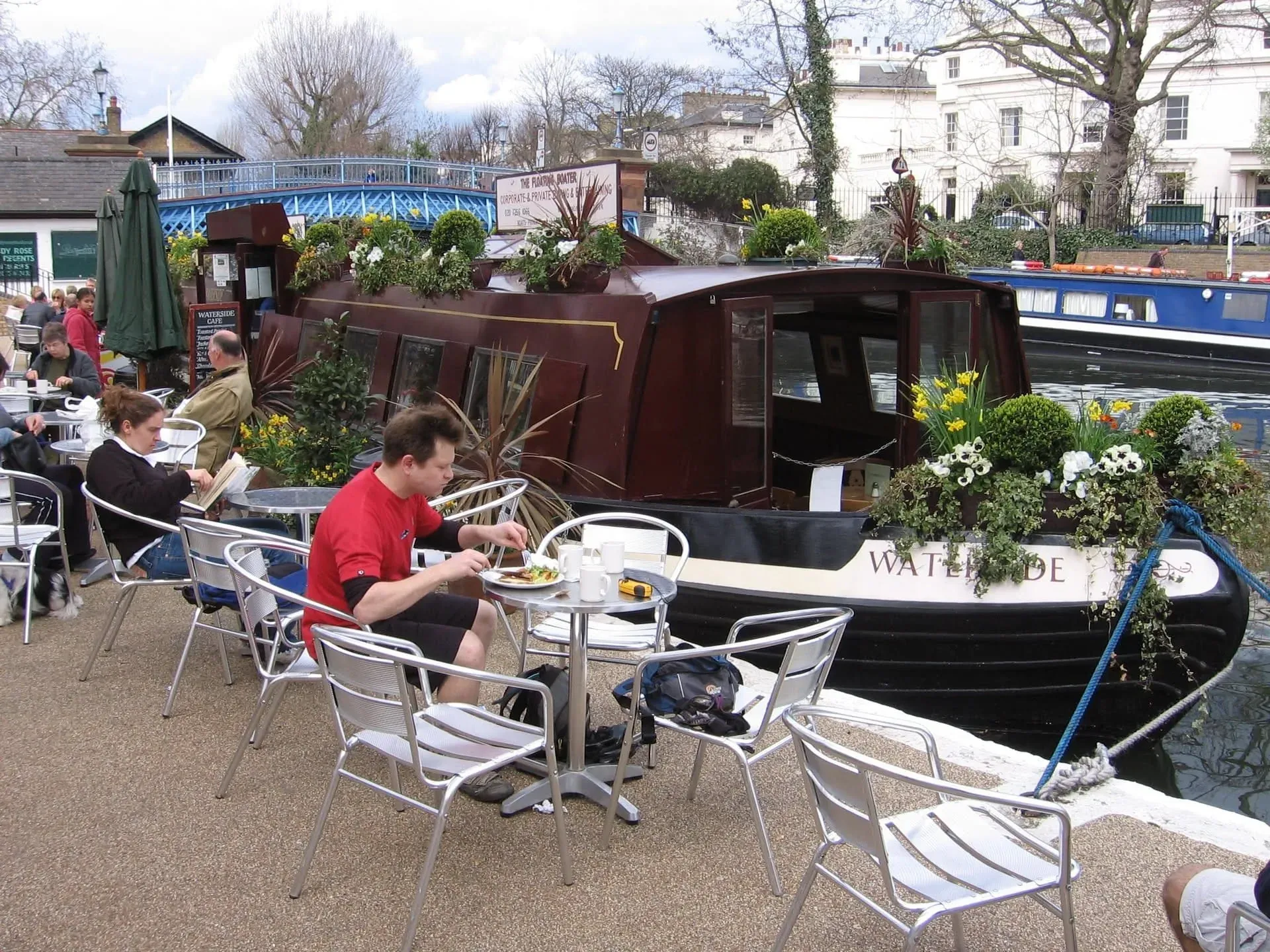Waterside cafe with diners at tables next to dark narrowboat on canal