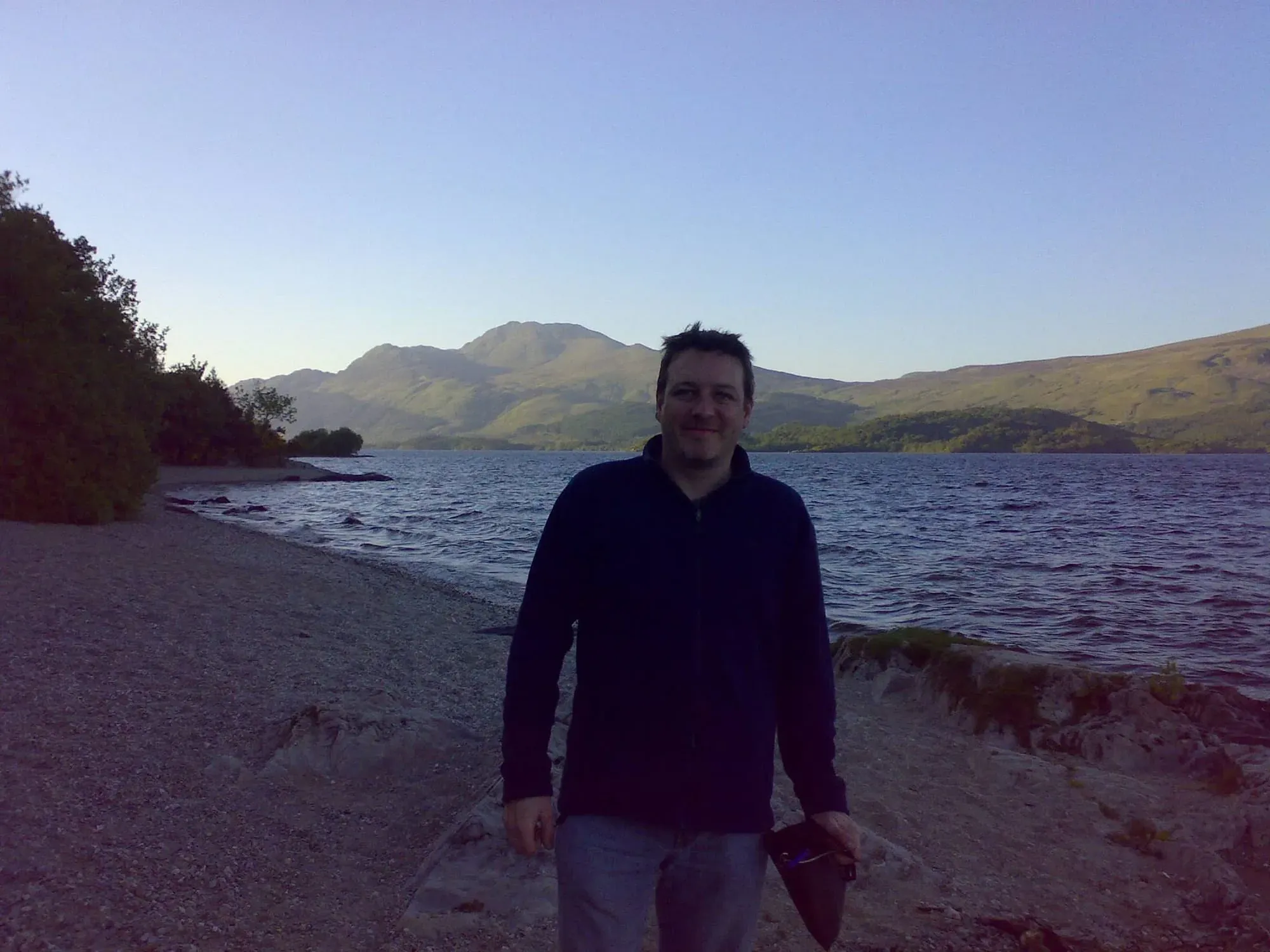 Man in black hoodie standing on beach with mountains and lake behind