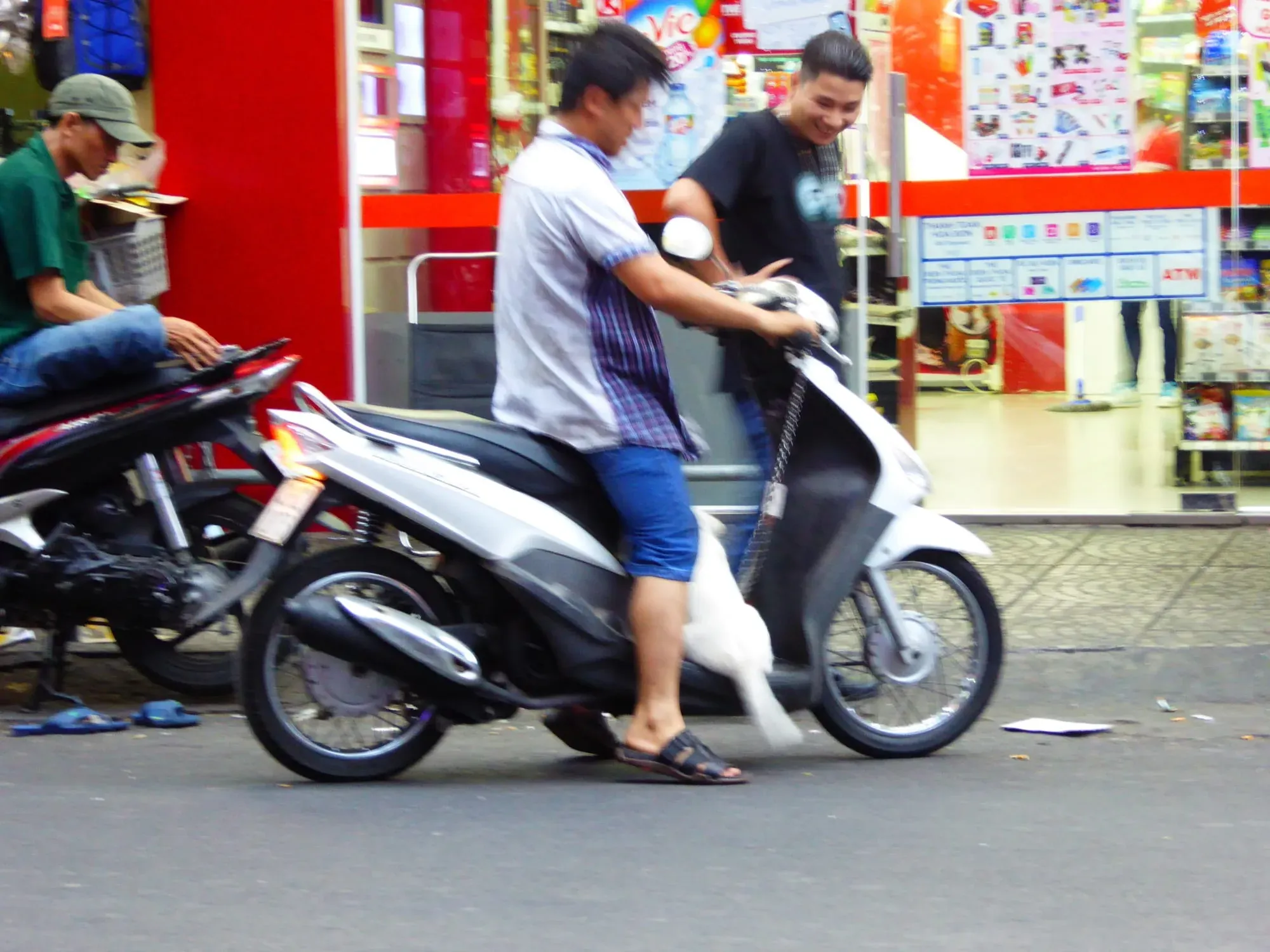 Two men on white and black motorbike on street with colorful shop signs