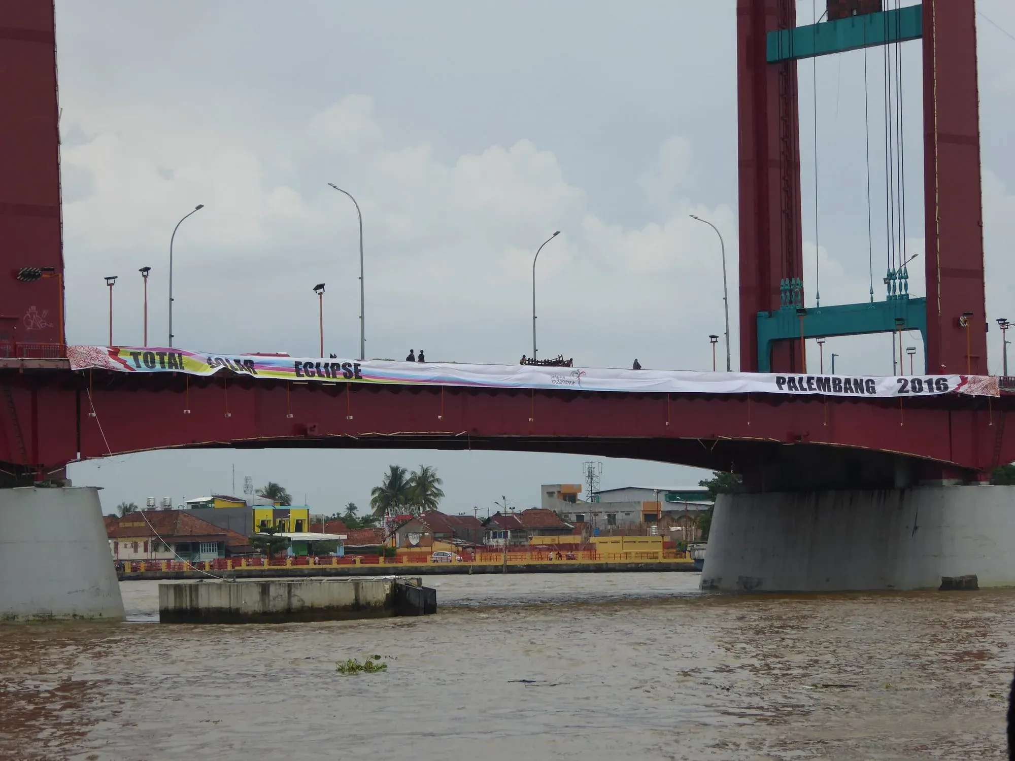 Red bridge with Palembang 2016 banner spanning muddy river with buildings below