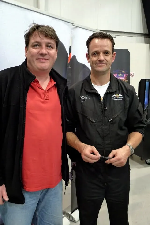 Two men standing together indoors, one in red shirt one in black