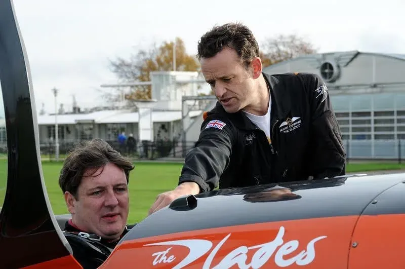 Man in black jacket gesturing toward red aircraft with hangar
