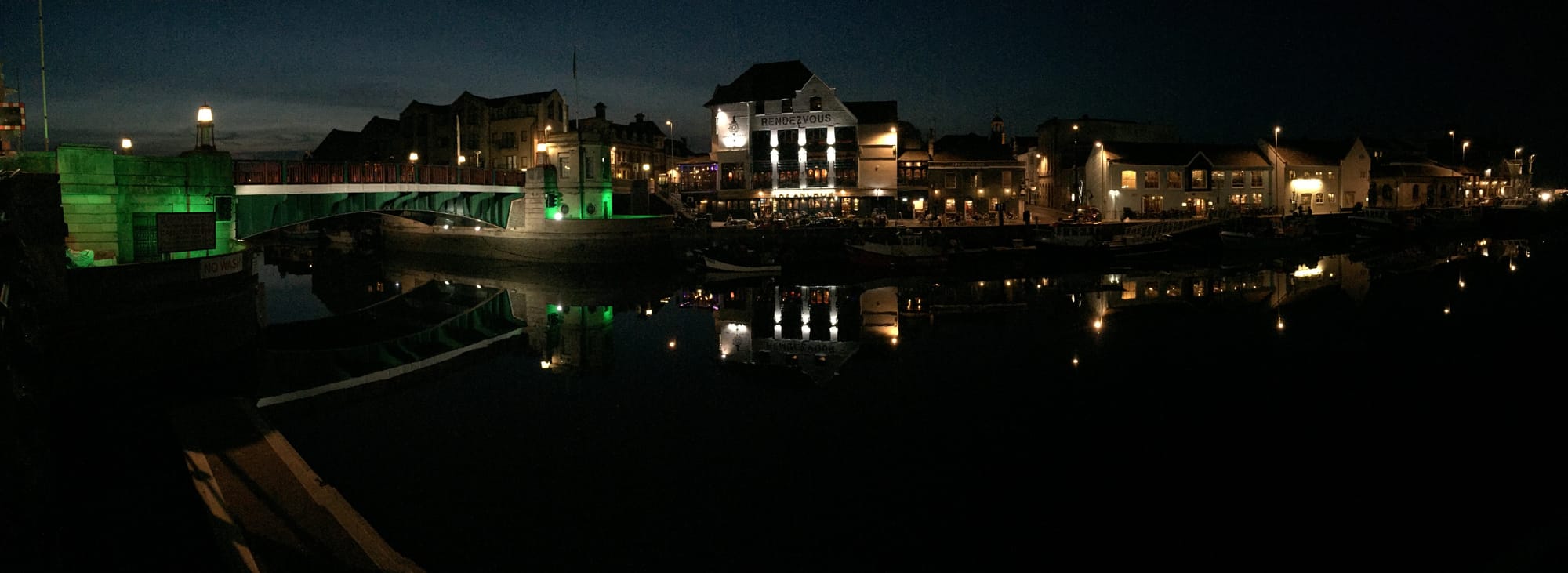 Night view of Weymouth harbor in Rodwell with green bridge and lit buildings reflected in water