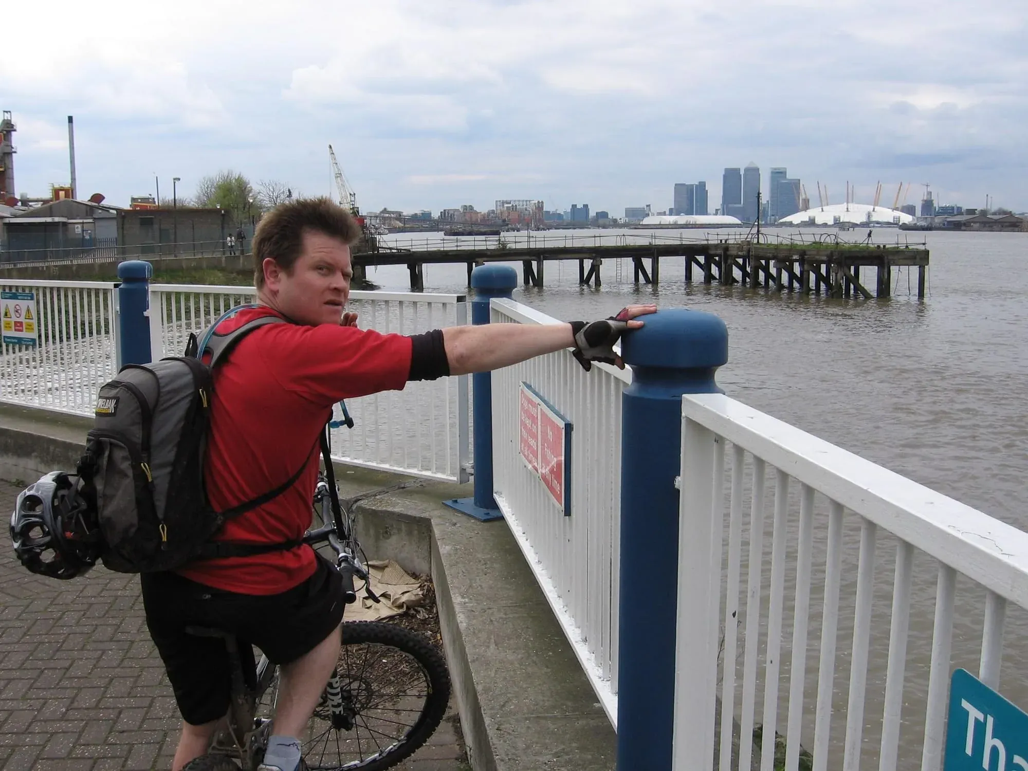 Cyclist in red jacket pointing across Thames toward Canary Wharf and Millennium Dome