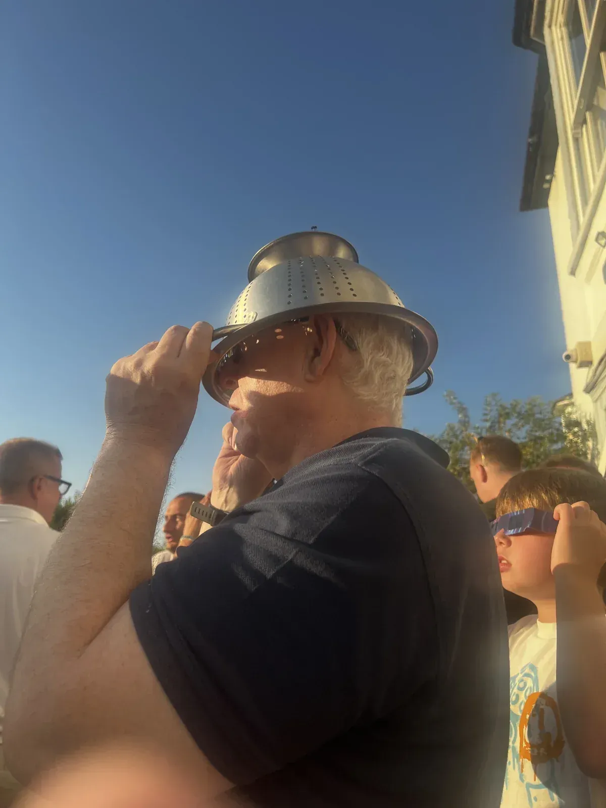 A man wearing a metal colander over his face as an improvised (and incorrect) eclipse viewer, a child beside him in proper eclipse glasses.