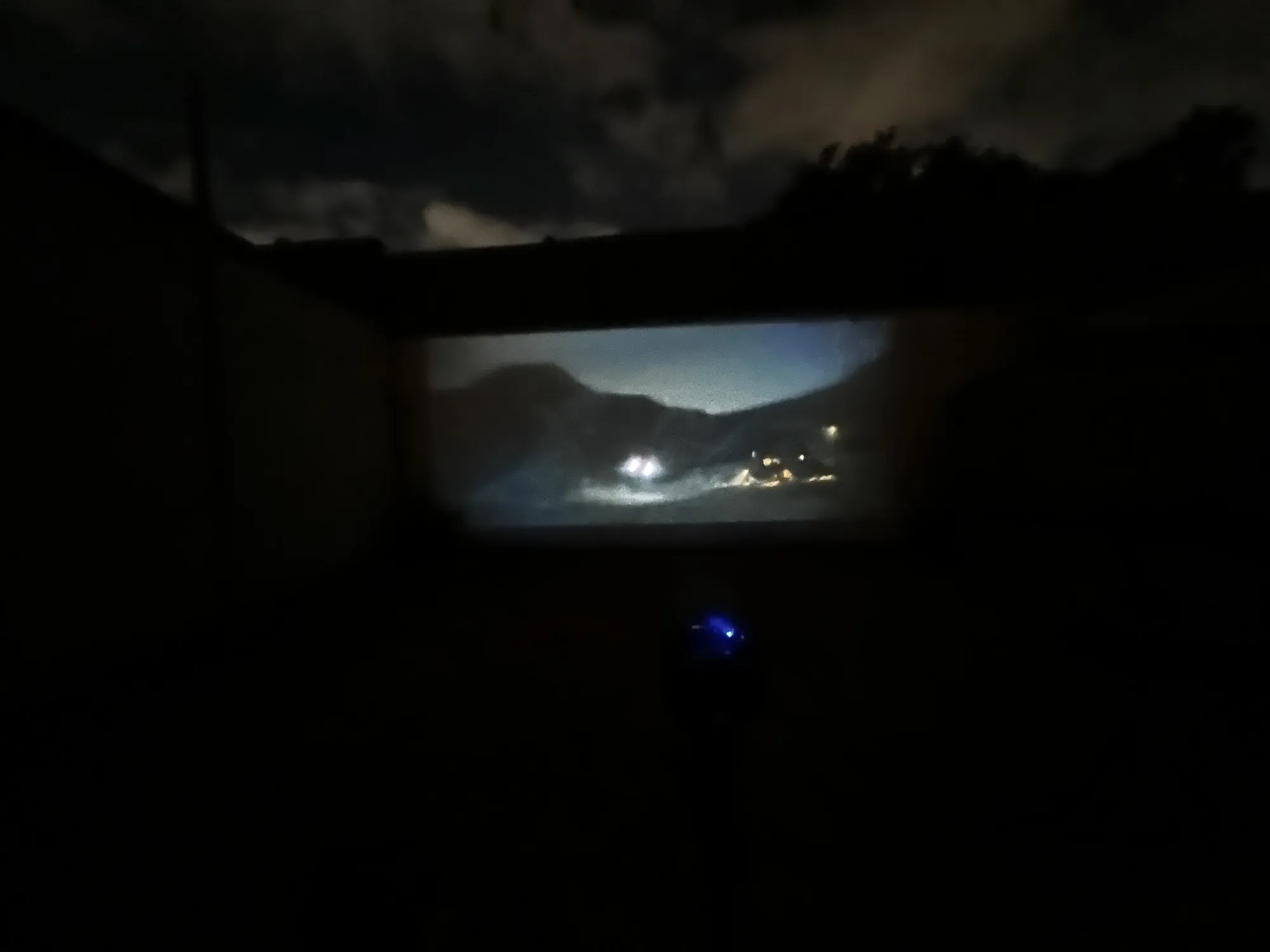 A film projected onto a garden wall at night showing a moonlit mountain village, the projector's blue light glowing below.